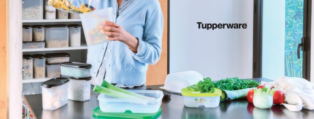 A bright kitchen scene showing a person using Tupperware storage containers while prepping vegetables, with a pantry of organized containers in the background—highlighting the everyday tools that help keep Cozy Chaos running smoothly.